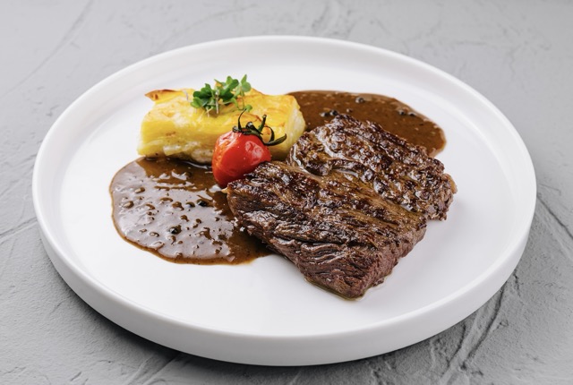 This close-up shot showcases a delectable grilled steak served with potato gratin and a vibrant red tomato on a clean white plate. The steak boasts a rich, brown crust, indicative of expertly searing the meat. The dish is garnished with a savory brown sauce, adding to its appeal. This image captures the essence of fine dining. This photo could be used for websites, brochures, menus, or advertisements related to the culinary arts, fine dining, restaurants, and food blogs. It communicates high-quality cuisine and a sophisticated dining experience.